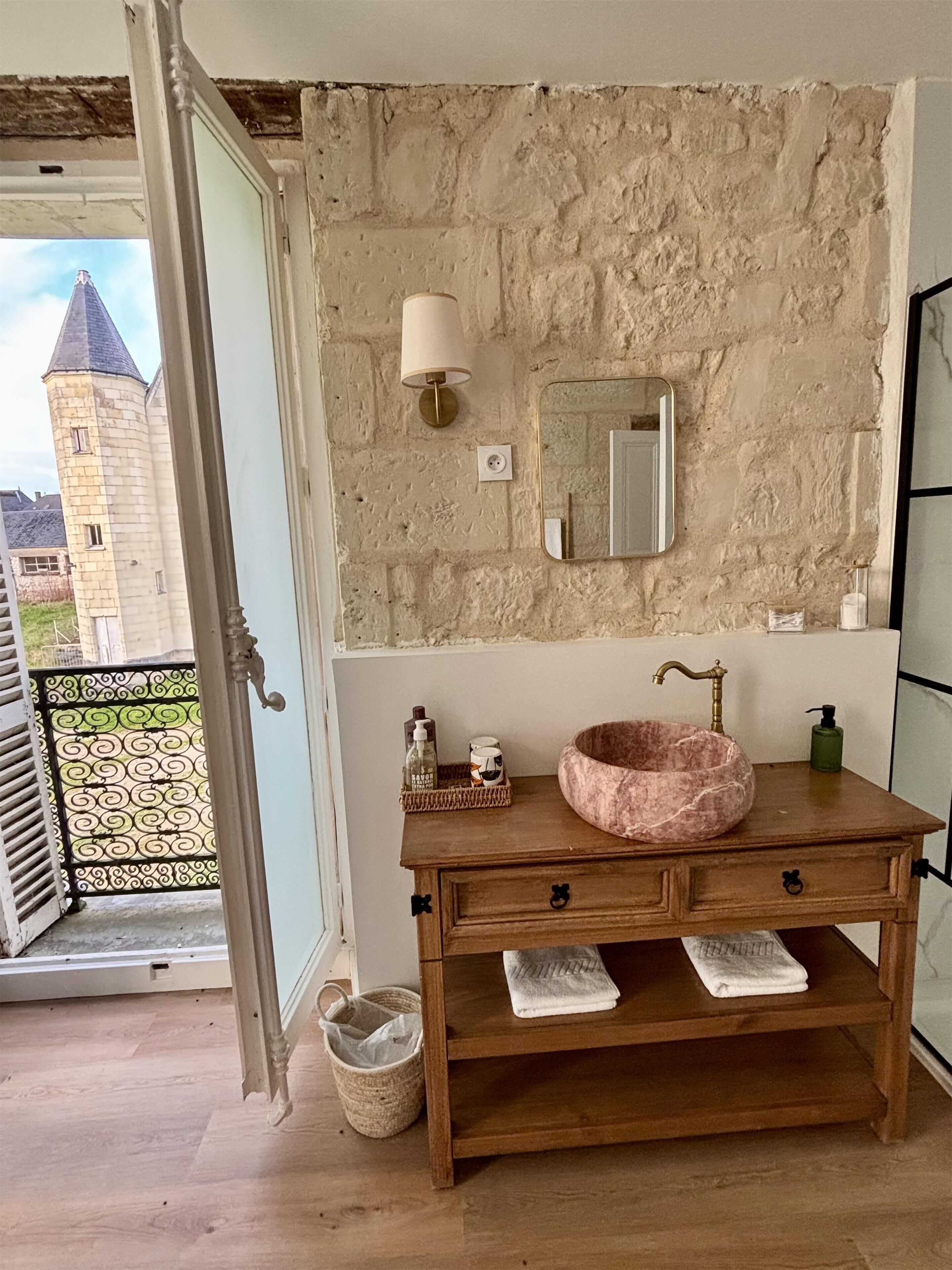 Een badkamer met een stenen muur, een houten wastafelmeubel, een marmeren wastafel, een spiegel, een wandlamp en uitzicht op een kasteel door een open deur.
