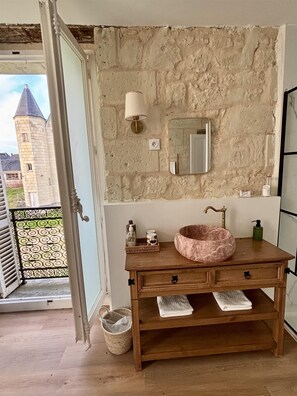 A bathroom with a stone wall, a wooden vanity, a marble sink, a mirror, a wall-mounted light, and a view of a castle through an open door.