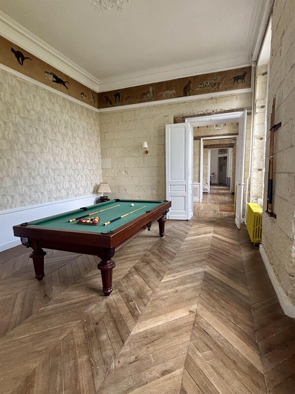 Une table de billard se trouve dans une pièce dont le sol est recouvert d’un parquet en chevrons, les murs sont blancs et une porte donne accès à une autre pièce.