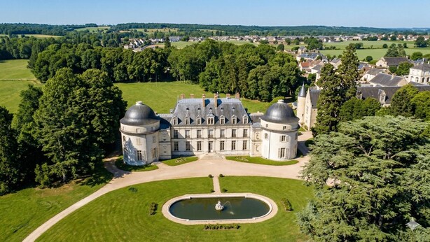 Un grand bâtiment historique, doté de deux imposants dômes, entouré d’une végétation luxuriante et d’une pelouse bien entretenue.