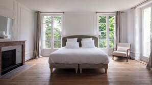 Une chambre avec un grand lit, un fauteuil, une cheminée et de grandes fenêtres.