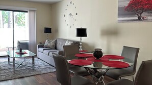 A modern living room with a dining area, featuring a glass table with red placemats, dark leather chairs, a grey sofa, and a wall clock. - Home away from home (Conway)