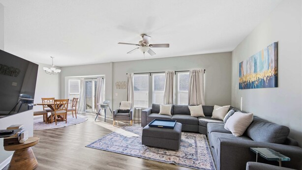 A modern living room with a grey sofa, ottoman, and a flat-screen TV. There is a dining area with a wooden table and chairs, and a ceiling fan. - Ark & Creation Retreat For 12! (Walton)