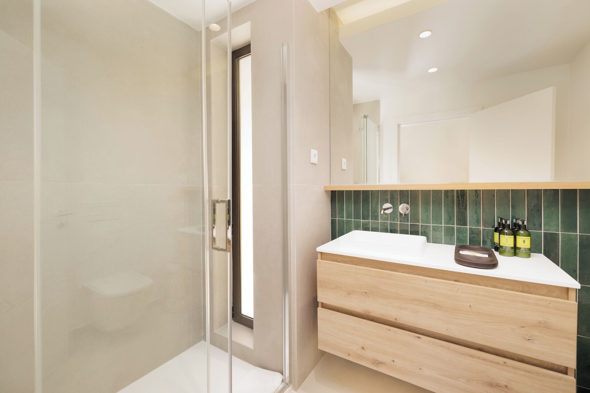 A modern bathroom with a glass shower enclosure, a white sink with a wooden vanity, and a green-tiled backsplash.