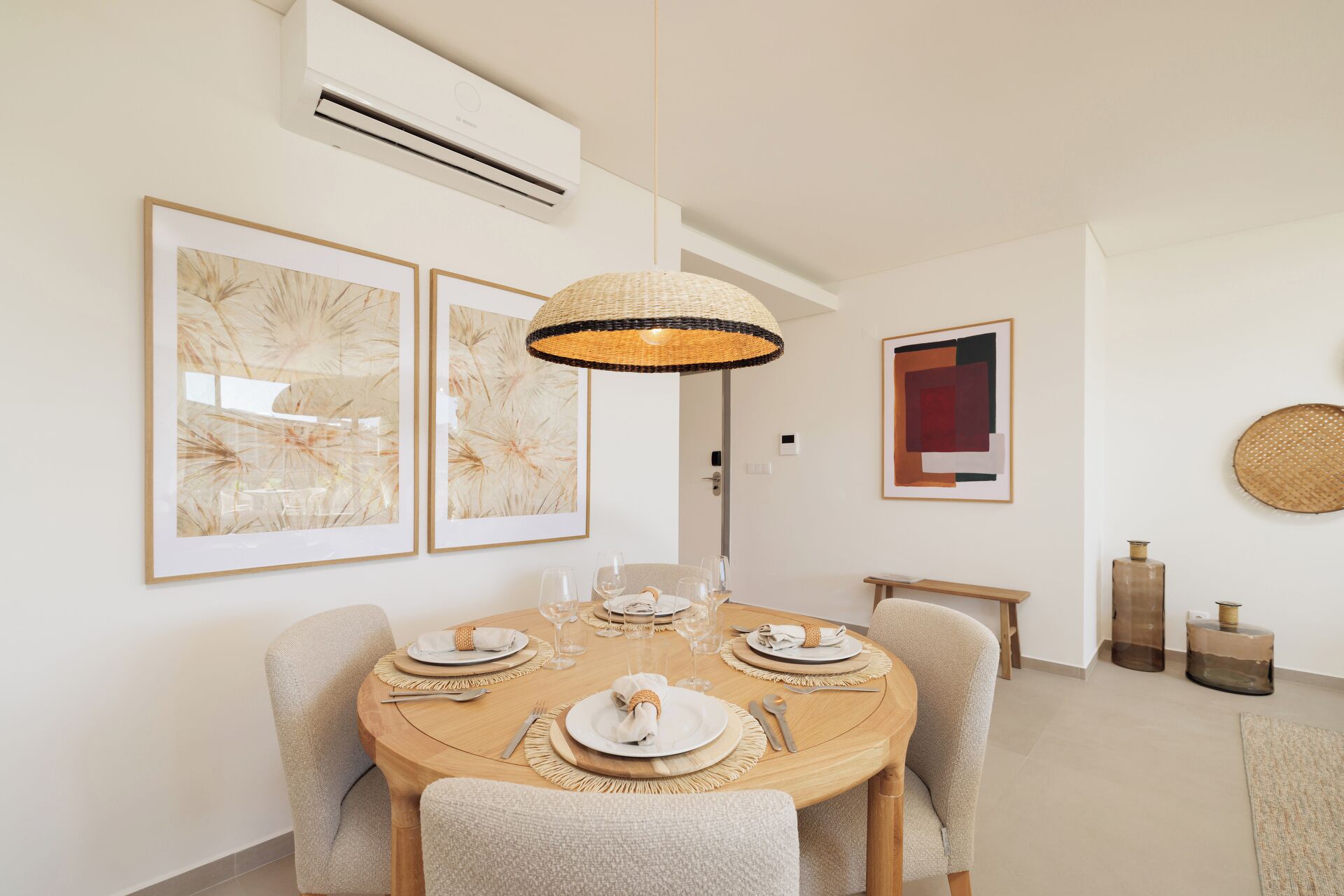 Een eetruimte met een ronde tafel voor vier personen, omringd door gestoffeerde stoelen. Aan de muur hangen twee abstracte schilderijen in lijst, een modern hanglamp en een klein bijzettafeltje met een kaars.