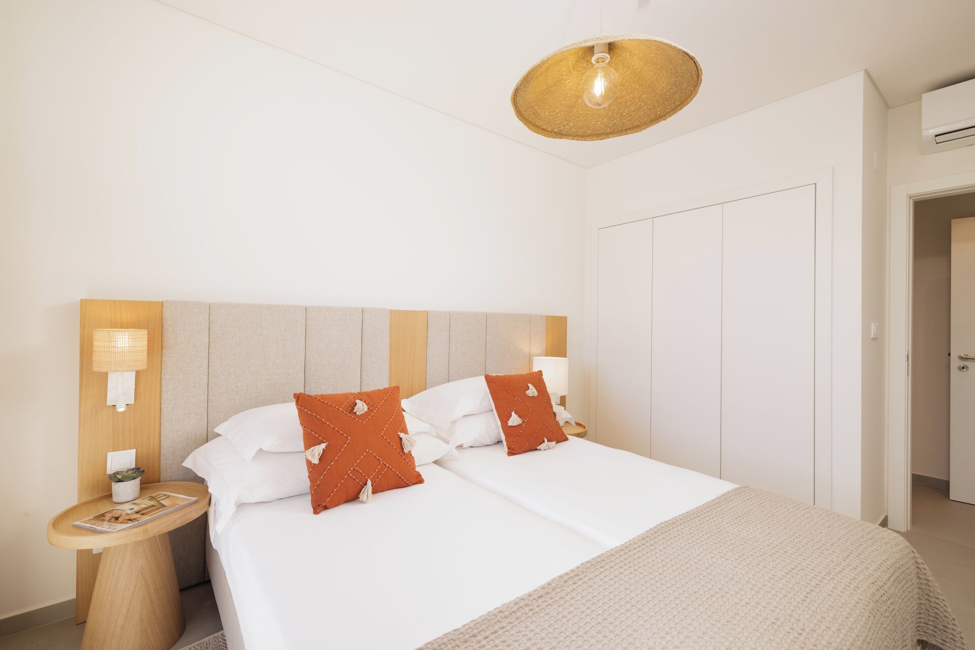 Quarto de hotel com uma cama, duas luminárias de cabeceira, um candeeiro tecido redondo, uma mesa de cabeceira de madeira e uma porta.