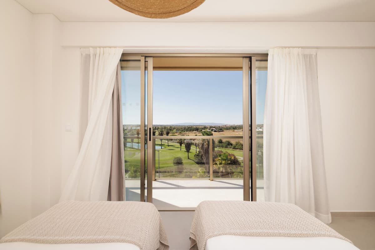 A bedroom with two beds, a large window with curtains, and a view of a golf course.