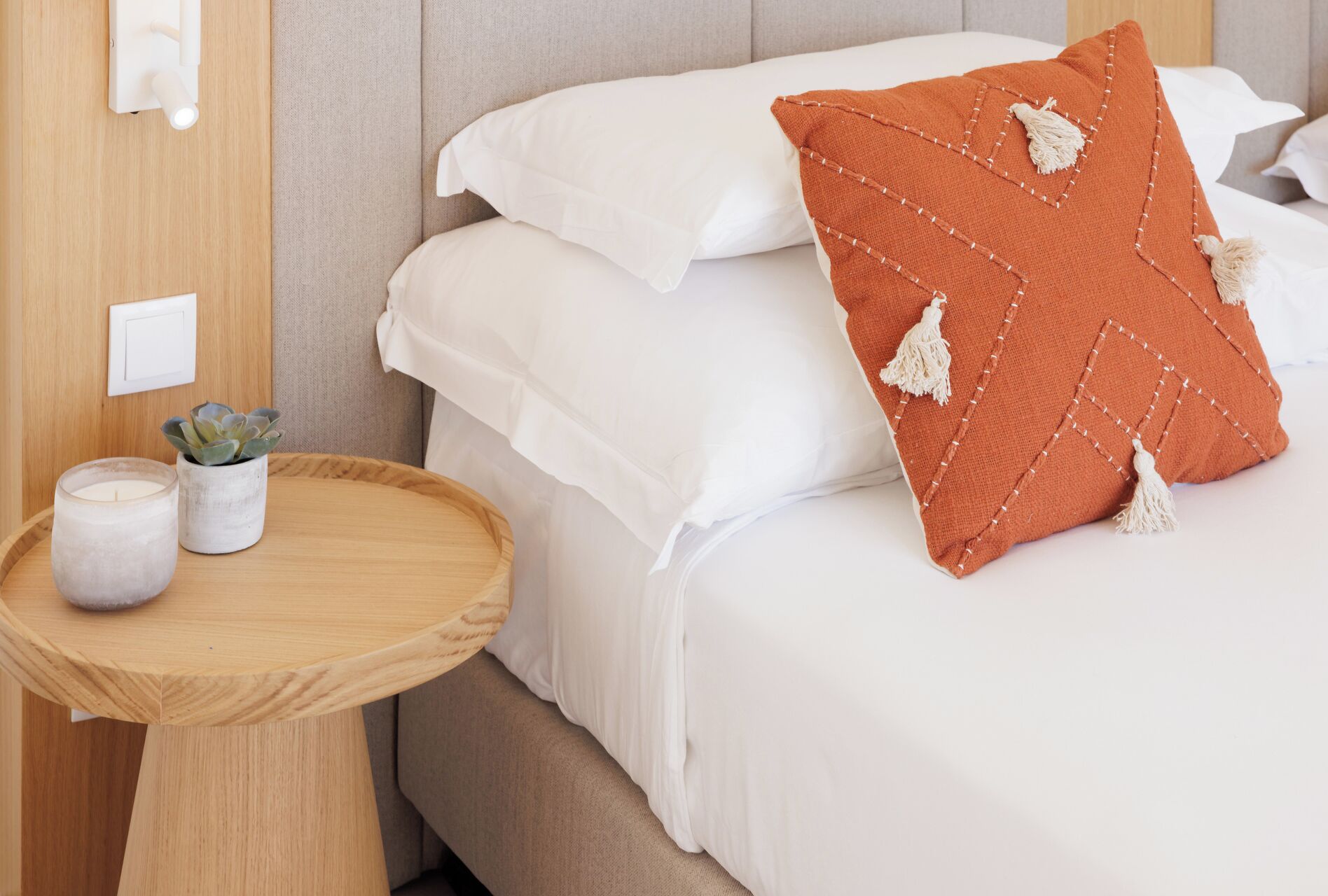 Een net opgemaakt bed met witte beddengoed en een decoratief oranje kussen. Een klein houten nachtkastje met een kaars en een potplantje.