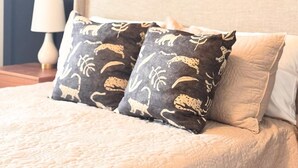 A bed with a beige quilt, two patterned pillows, a white pillow, and a bedside lamp. - The Adelyn, where comfort meets convenience (Jeffersonville)