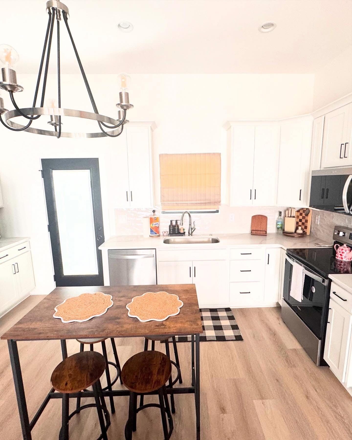 A modern kitchen with white cabinets, a black countertop, a wooden dining table with two chairs, a chandelier, and a window with blinds.
