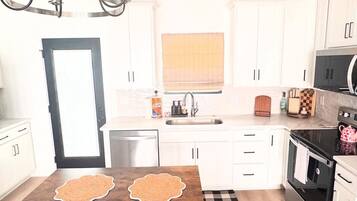 A modern kitchen with white cabinets, a black countertop, a wooden dining table with two chairs, a chandelier, and a window with blinds.