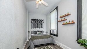 Ein Schlafzimmer mit einem Bett, einem karierten Teppich, einem Deckenventilator und Regalen an der Wand mit Pflanzen.