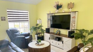 Een woonkamer met een grote flatscreen televisie, leren banken, een bijzettafeltje en potplanten.