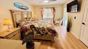 Een slaapkamer met een bed, nachtkastje, lamp, plafondventilator, televisie, raam met gordijnen, stoel en een potplant.
