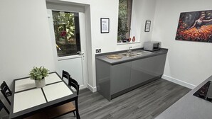 A modern kitchen with a dining area, featuring a large island, grey cabinets, and a window with a view of greenery. - Shawforth - 2 Beds - Sleeps 4 - Street Parking (Rochdale)
