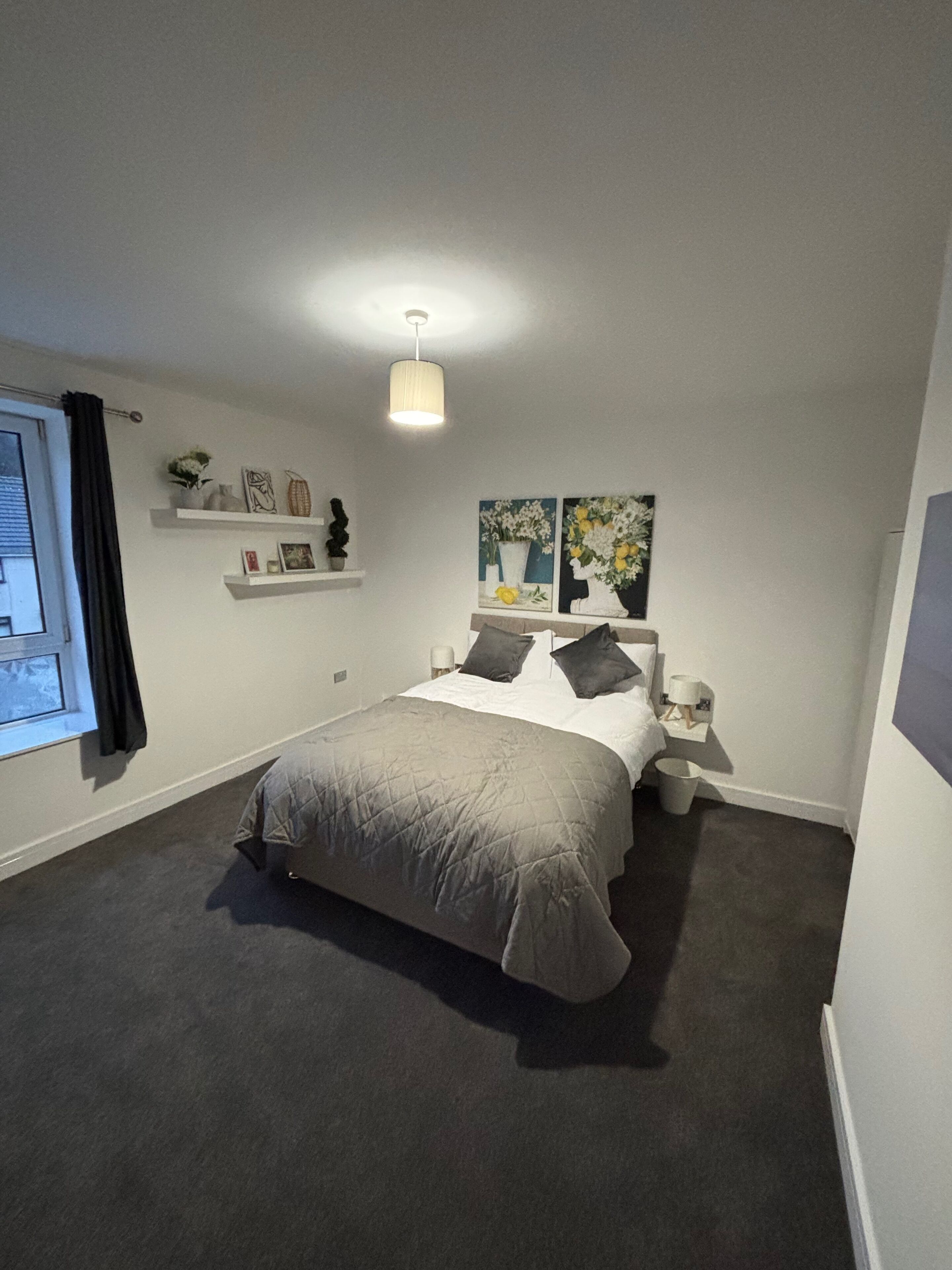 A bedroom with a bed, bedside tables, a window with curtains, and wall-mounted shelves with decorative items.