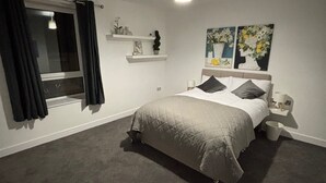 A bedroom with a bed, a nightstand, a window with curtains, a ceiling light, and wall-mounted shelves with decorative items. - Shawforth - 2 Beds - Sleeps 4 - Street Parking (Rochdale)