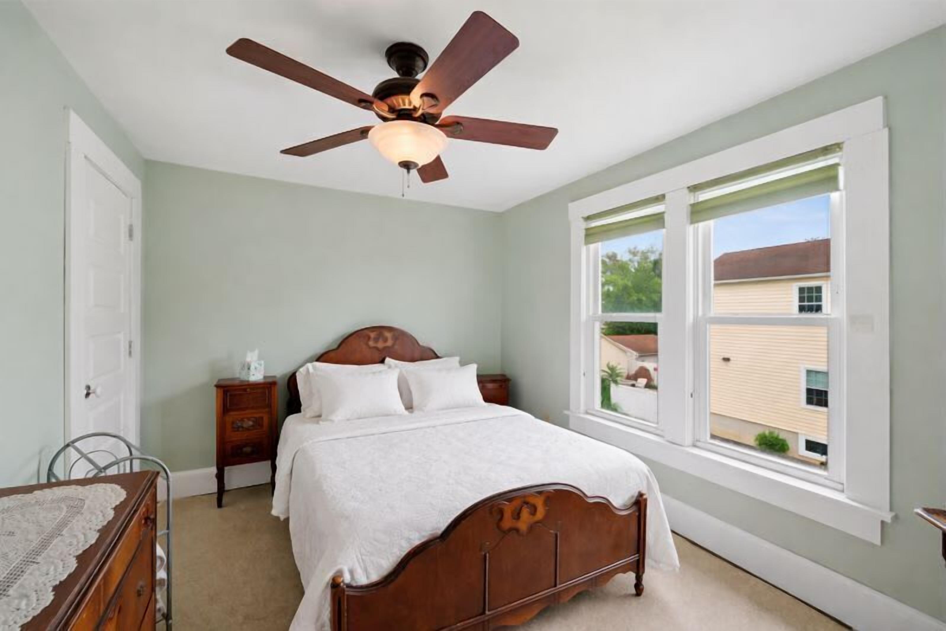 A bedroom with a wooden bed, a ceiling fan, a nightstand, and a window with a view of a house.