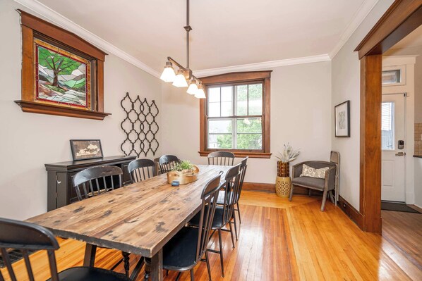 Un comedor con mesa y sillas de madera, una araña y una ventana con vista a follaje.