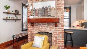 A living room with a brick fireplace, a yellow armchair, a bench, and a kitchen area with a bar stool. - Historic Retreat Gateway (Dayton)