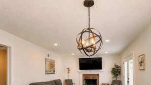 A dining area with a wooden table and chairs, a gray sofa, a TV mounted above a fireplace, and a chandelier. - Lark Meadow Retreat Sleeps 10! (Mason)
