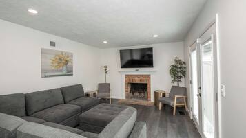 A modern living room with a grey sectional sofa, a fireplace, two armchairs, a flat-screen TV mounted above the fireplace, and a potted plant.
