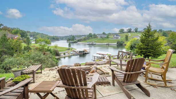 A lakeside patio with wooden Adirondack chairs and a fire pit, overlooking a calm lake and a dock area. - Lazy Days Waterfront for 12! (Williamstown)