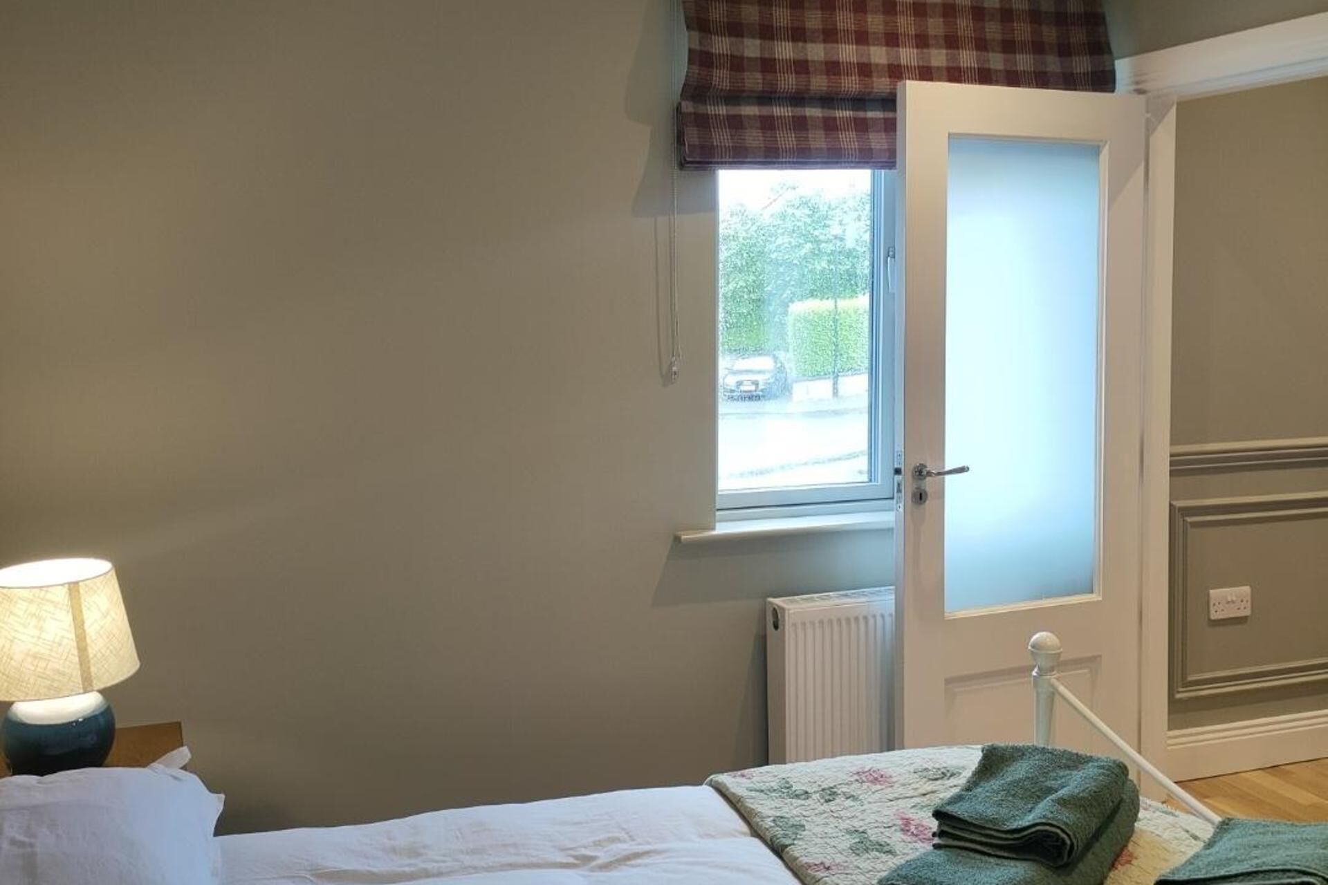 A bedroom with a bed, a window with a view of the outside, a radiator, and a lamp on a bedside table.