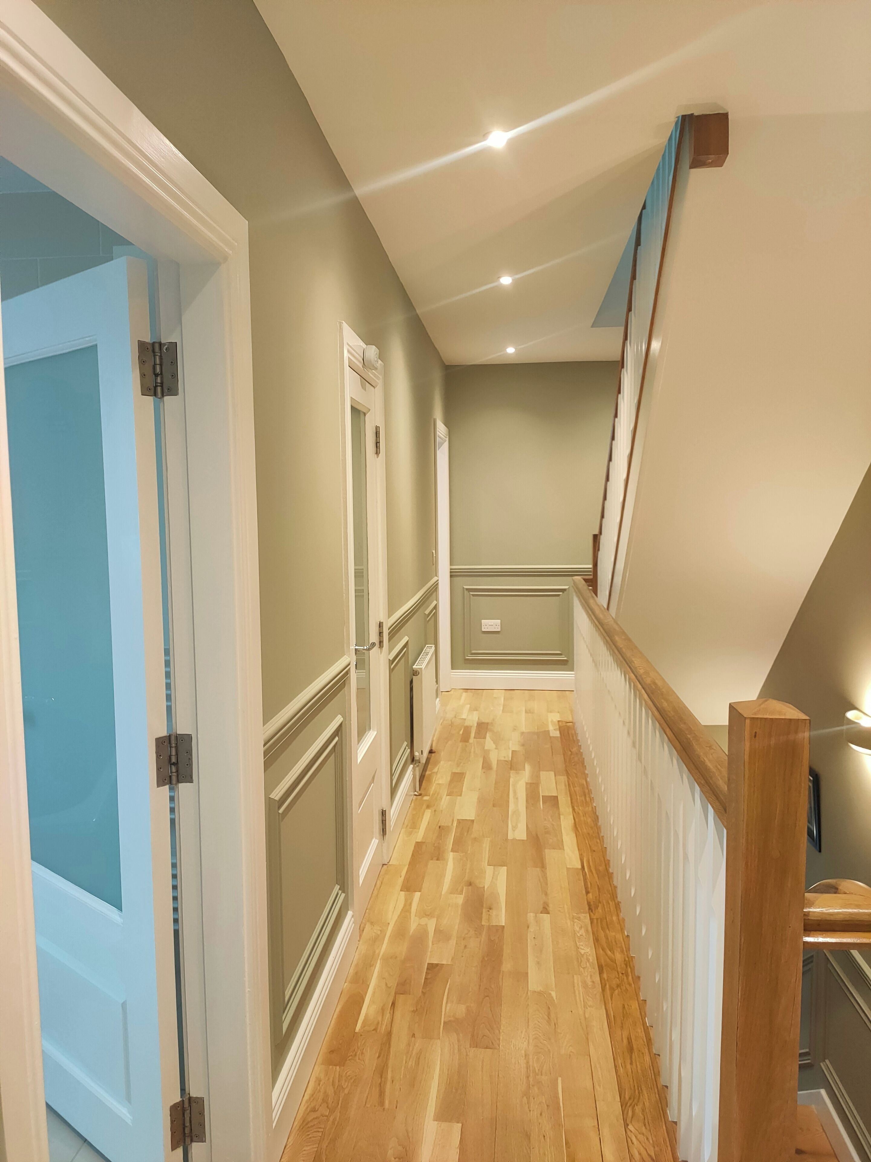 A hallway with wooden flooring, a blue door on the left, and a staircase with a wooden railing on the right.
