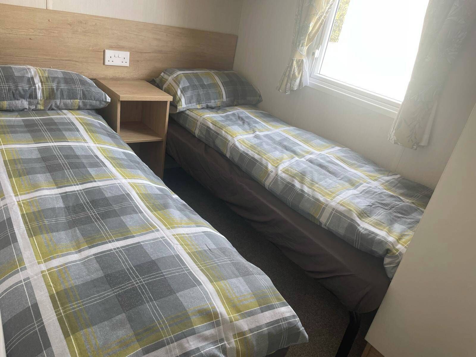 Een studentenkamer met twee eenpersoonsbedden, een houten nachtkastje en een raam met gordijnen.