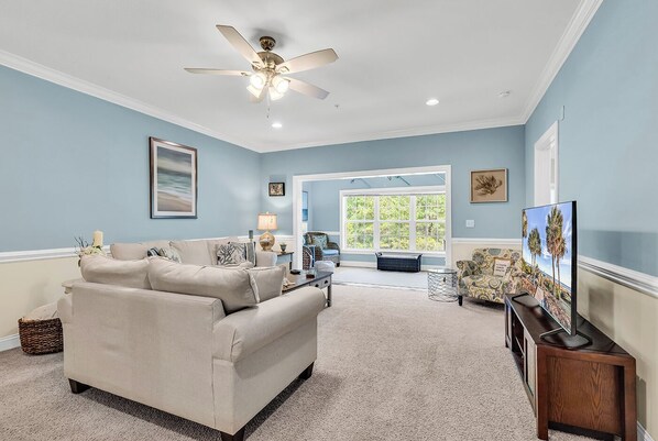 A living room with a beige sofa, a ceiling fan, a large window, and a television.