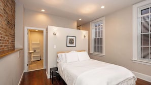 A bedroom with a wooden floor, a white bed, a door leading to a bathroom, and a window with blinds. - Loft Life Pendleton 2 Chic Downtown Condo for 4! (Cincinnati)