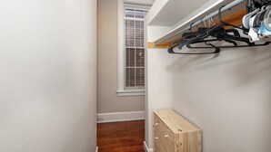 A narrow hallway with a wooden floor, a window with blinds, and a built-in shelving unit with hanging items. - Loft Life Pendleton 2 Chic Downtown Condo for 4! (Cincinnati)