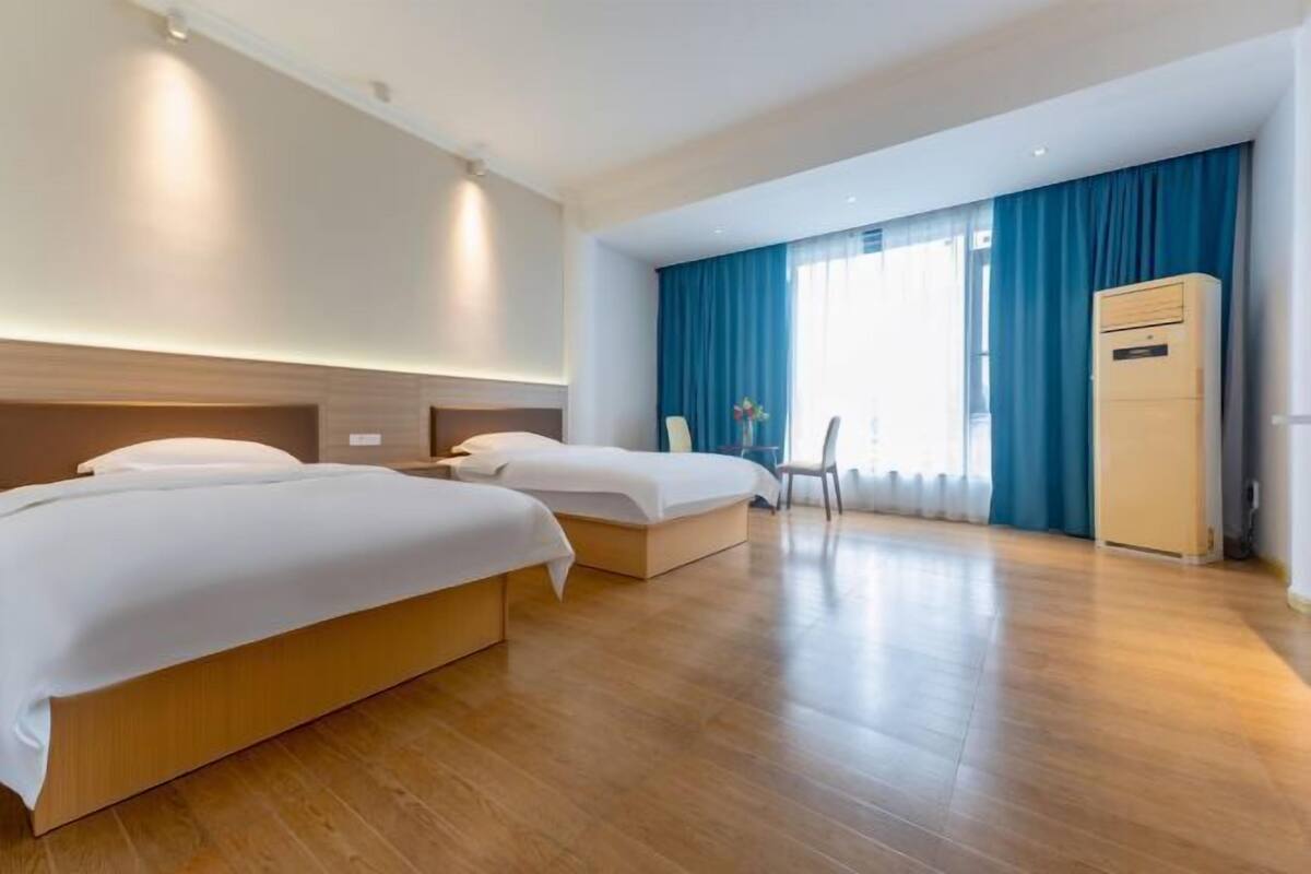 Habitación de hotel con dos camas, piso de madera, un ventanal grande con cortinas azules y un aire acondicionado de pared.