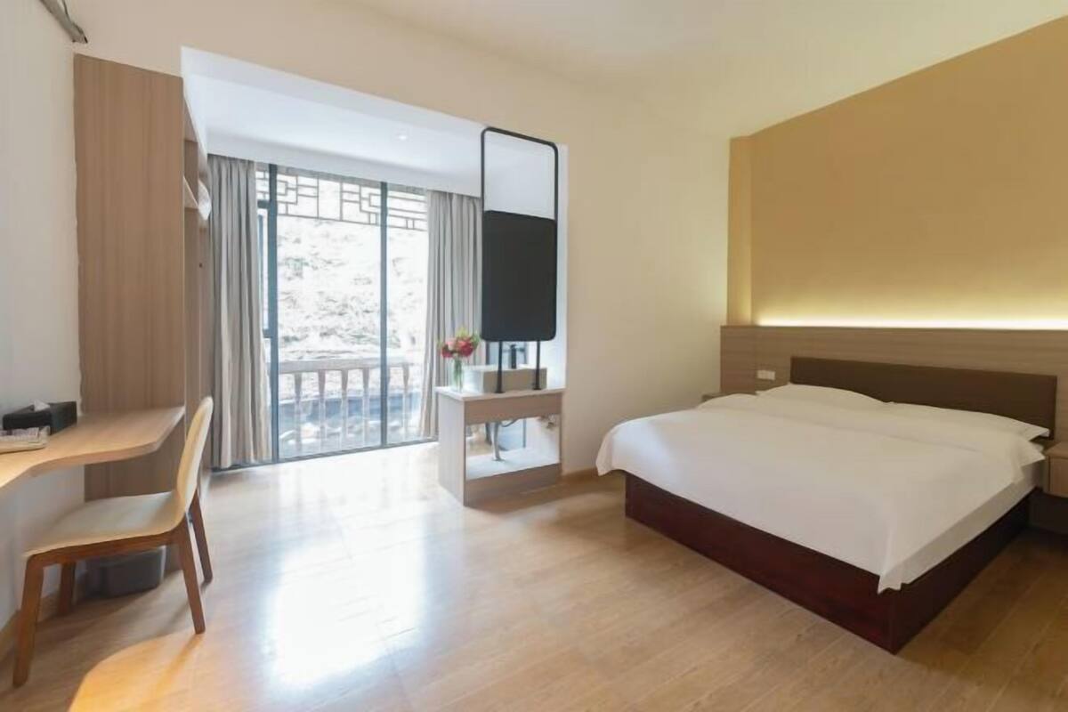 Habitación de hotel con cama, escritorio, silla, televisor y balcón con vista a árboles.