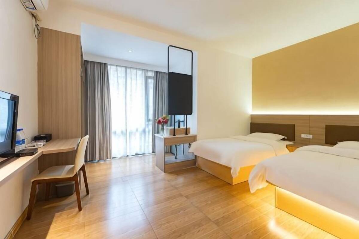 Habitación de hotel con escritorio, dos camas, televisor y un ventanal grande con cortinas.
