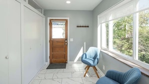 A hallway with a wooden door, a blue chair, and a window with a view of greenery. - Love n Life Haven (Cincinnati)