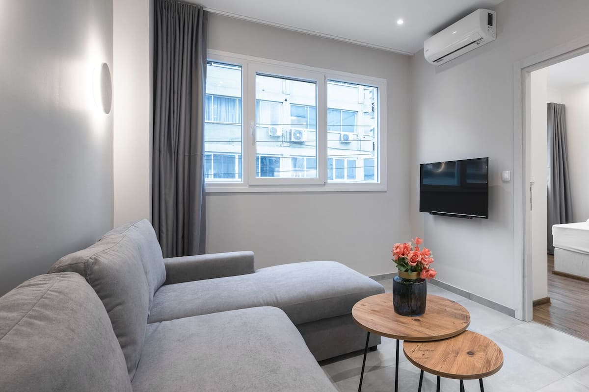 Een moderne woonkamer met een grijze bank, een ronde houten salontafel met een bloevas, een flatscreen-televisie aan de muur en een raam met grijze gordijnen.