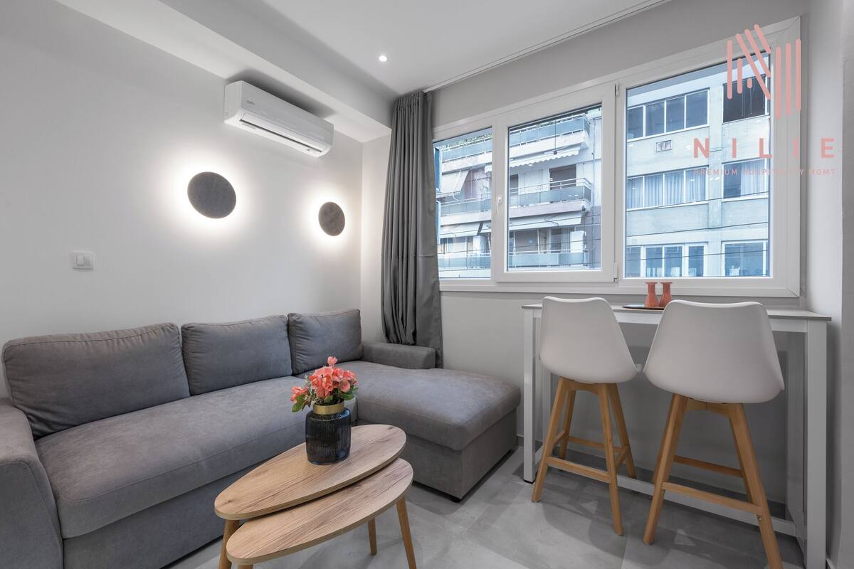 Een moderne woonkamer met een grijze bank, een klein rond tafeltje met een vaas, twee barkrukken en een raam met uitzicht op gebouwen.