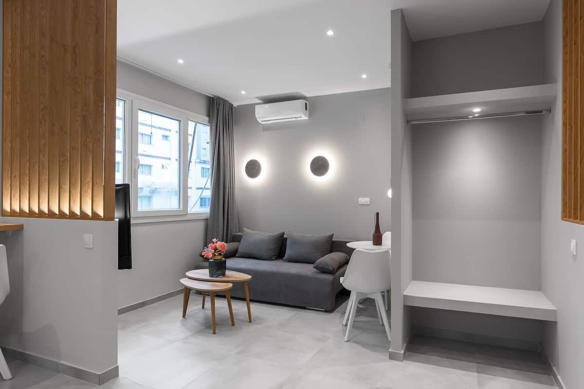 Een moderne woonkamer met een grijze bank, een klein rond tafeltje met een vaas, en een witte stoel. Er is een inbouwkbank met een witte tafel, en een airconditioner aan de muur.