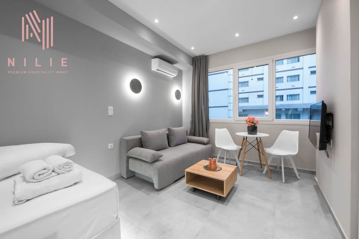 Een moderne hotelkamer met een grijze bank, een wit bed, een klein houten bijzettafeltje en een aan de muur bevestigde televisie.