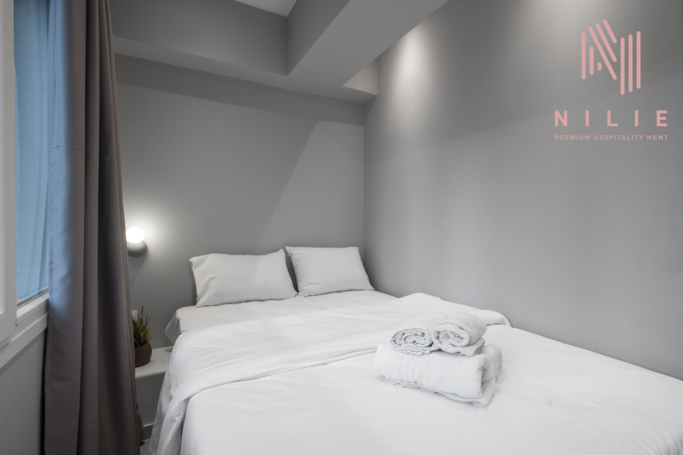 Una cama bien tendida con sábanas blancas y toallas enrolladas, una planta en la mesita de noche y un aplique de pared en una habitación moderna y minimalista.