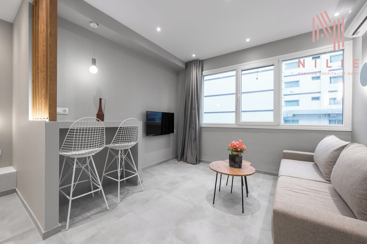 Een moderne woonkamer met een bank, twee open stoelen, een klein rond tafeltje met een bloevas, een aan de muur bevestigde televisie en een raam met gordijnen.
