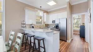 A modern kitchen with white cabinets, a stainless steel refrigerator, a built-in microwave, and a granite countertop. There are three bar stools and a coffee maker on the counter. - Turquoise Tail (Panama City Beach)
