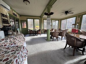 A dining area with wicker chairs, a table with a floral tablecloth, and a view of trees through large windows. - Inn at Lake Joseph (Forestburgh)