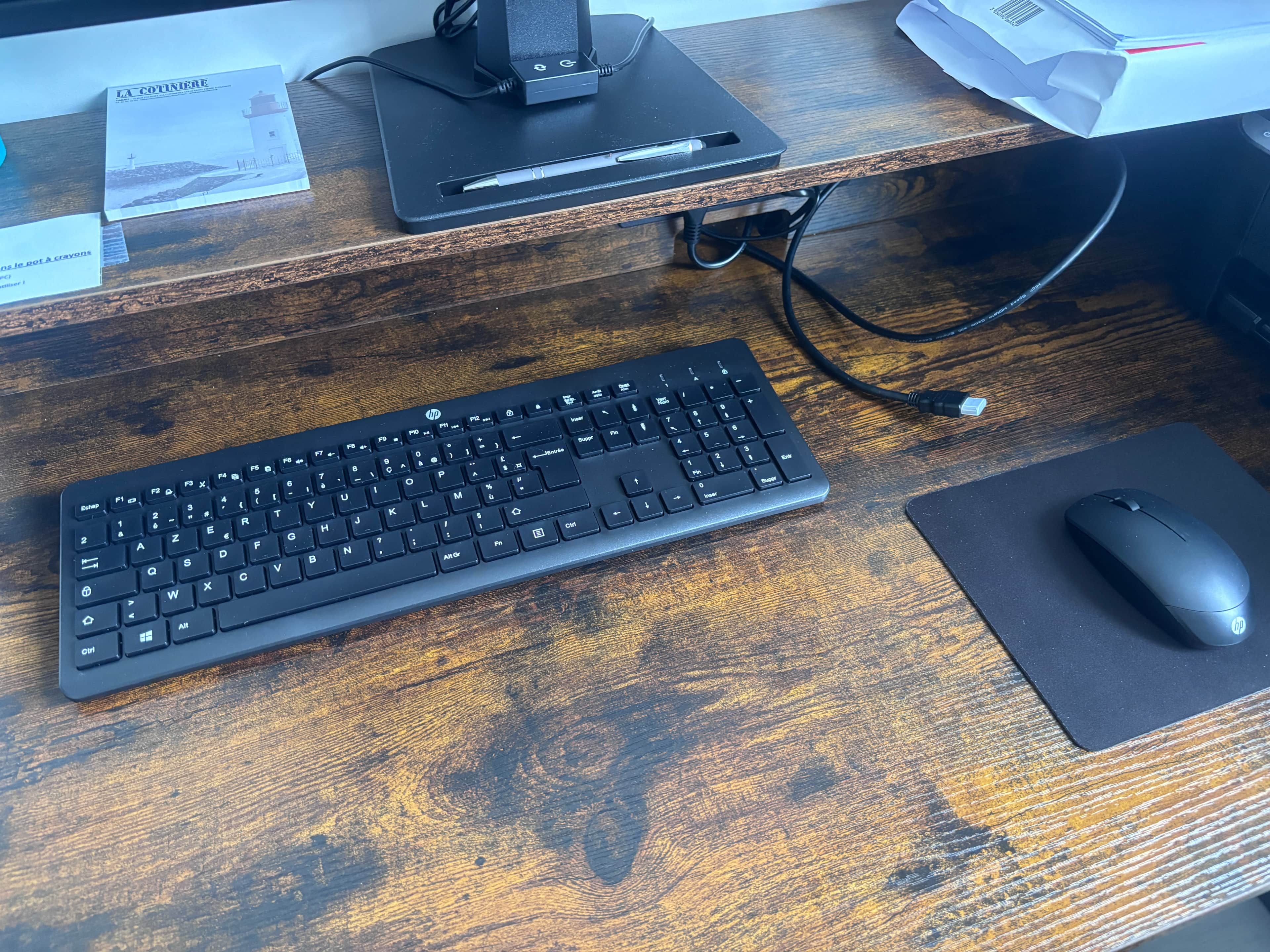 Ein Holztisch mit einer schwarzen HP-Tastatur, einer schwarzen HP-Maus und einem schwarzen Monitorständer.