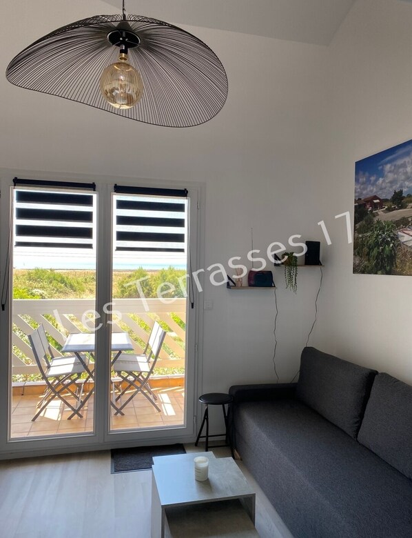 A modern living room with a grey sofa, a white coffee table, and a hanging light fixture. There is a sliding glass door leading to an outdoor patio with a view of greenery and a building.