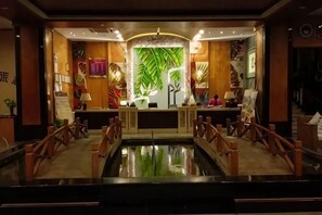 Un área de recepción con mostrador, una pared decorativa con hojas de palmera, una pequeña piscina y barandillas de madera.