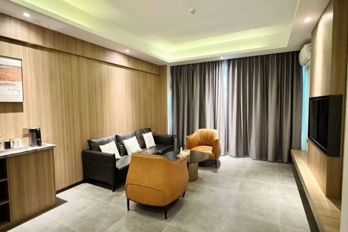 Ein modernes Wohnzimmer mit Sofa, Sessel und Couchtisch, einem an der Wand montierten Fernseher und einem fensterbedeckenden Vorhang.