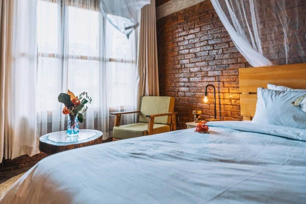 A hotel room with a large bed, a chair, a small table with a vase of flowers, and a brick wall.